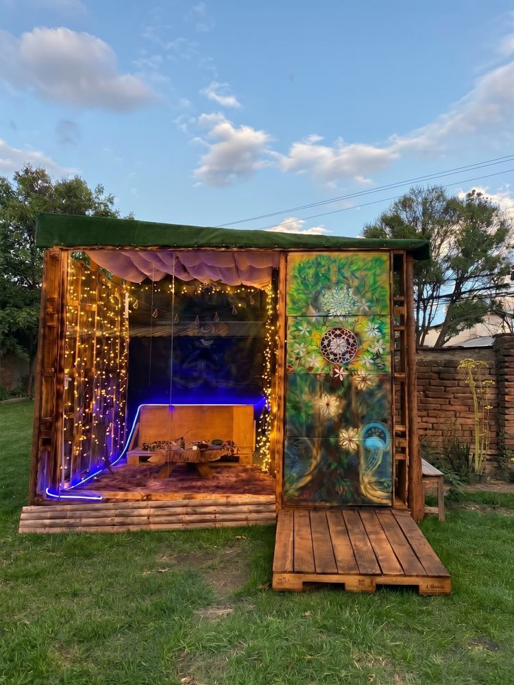 Casa del Oráculo, a painted wooden hut with fairy lights, surrounded by green grass