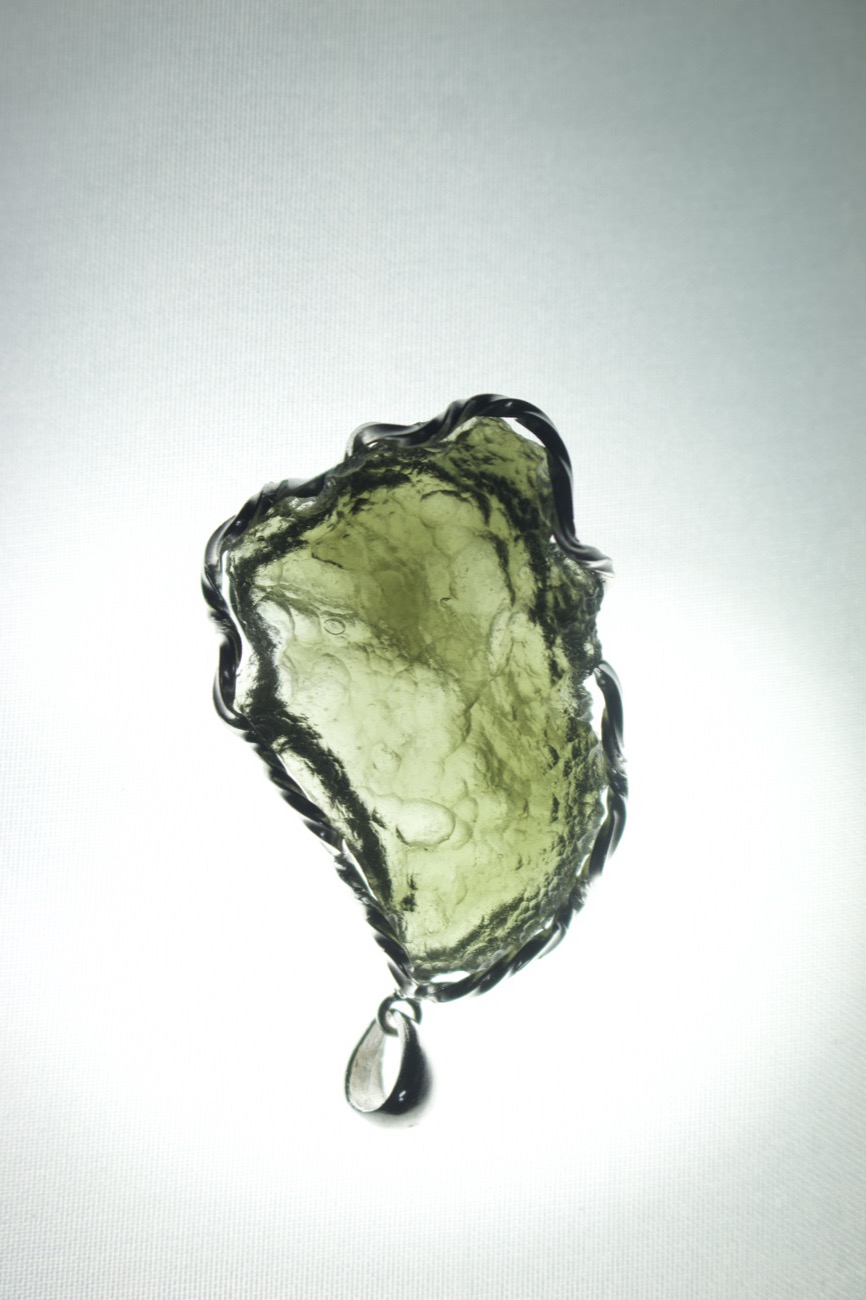 A translucent green Moldavite pendant wrapped in dark wire, lit from behind